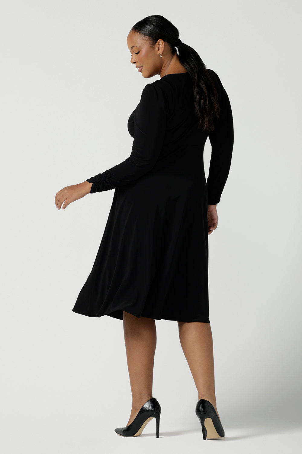 Back view of a size 16 woman wears a Alyssa Dress in jersey. V-neckline style great for work to weekend wear. Corporate dress for women. Travel friendly with an empire line style. Midi dress and styled back with black strappy heels. Made in Australia for women size 8 - 24.