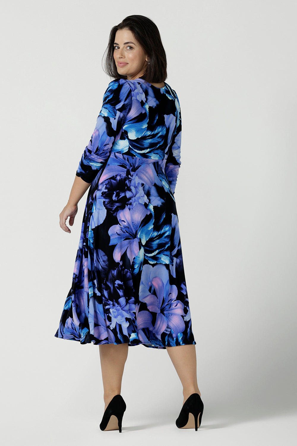 Back view of a Size 10 woman wears the Alyssa Dress in Blue Lily, Empire line style with bold blue flowers on a black base. Deep V - neckline and size inclusive. Made in Australia for women size 8 - 24. Petite to plus size women. Dresses for workwear to event dressing.
