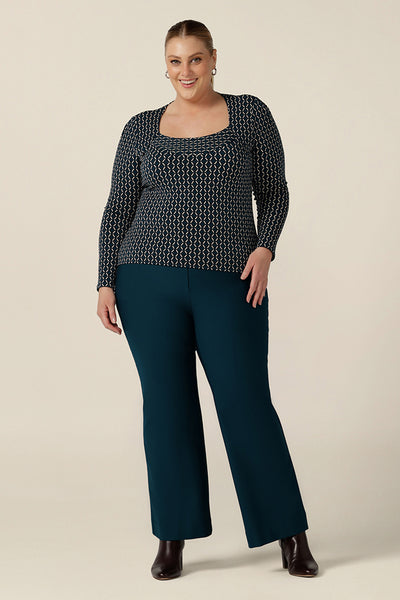 A plus size, size 18 woman wears a long sleeve top with squared scoop neckline. worn with petrol blue, bootcut tailored pants, the geometric print jersey top is comfortable for work wear capsule wardrobes. Made in Australia, shop tops in sizes 8 to 24.