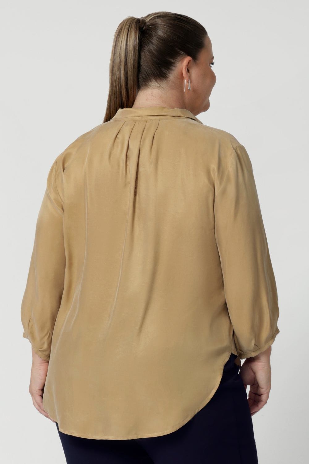 Size 18 woman wears golden coloured workwear blouse. Featuring a sharp collar neckline and tuck details below the back neckline, this blouse exudes an understated yet refined sophistication. The 3/4 length sleeves with narrow, bound hems add a touch of subtle detail. Ensuring woman's light weight blouse feel both modern and timeless. Available in sizes 8-24.