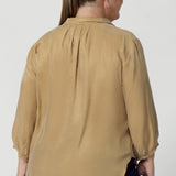 Size 18 woman wears golden coloured workwear blouse. Featuring a sharp collar neckline and tuck details below the back neckline, this blouse exudes an understated yet refined sophistication. The 3/4 length sleeves with narrow, bound hems add a touch of subtle detail. Ensuring woman's light weight blouse feel both modern and timeless. Available in sizes 8-24.