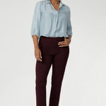 Semi-fitted through the body, it features a classic collar neckline and subtle tuck details below the back neckline, adding gentle structure without compromising comfort. Over 30s woman wears her light blue shirt tucked into burgundy coloured work pants.