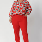 The semi-fitted silhouette ensures a flattering shape without being too tight, offering ease of movement for all-day wear. Below the back neckline, tuck details add an element of interest and elevated tailoring. Size 18 woman wear red printed blouse with tangerine coloured pants for a statement workwear outfit. 