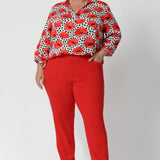 The semi-fitted silhouette ensures a flattering shape without being too tight, offering ease of movement for all-day wear. Below the back neckline, tuck details add an element of interest and elevated tailoring. Size 18 woman wear red printed blouse with tangerine coloured pants for a statement workwear outfit. 