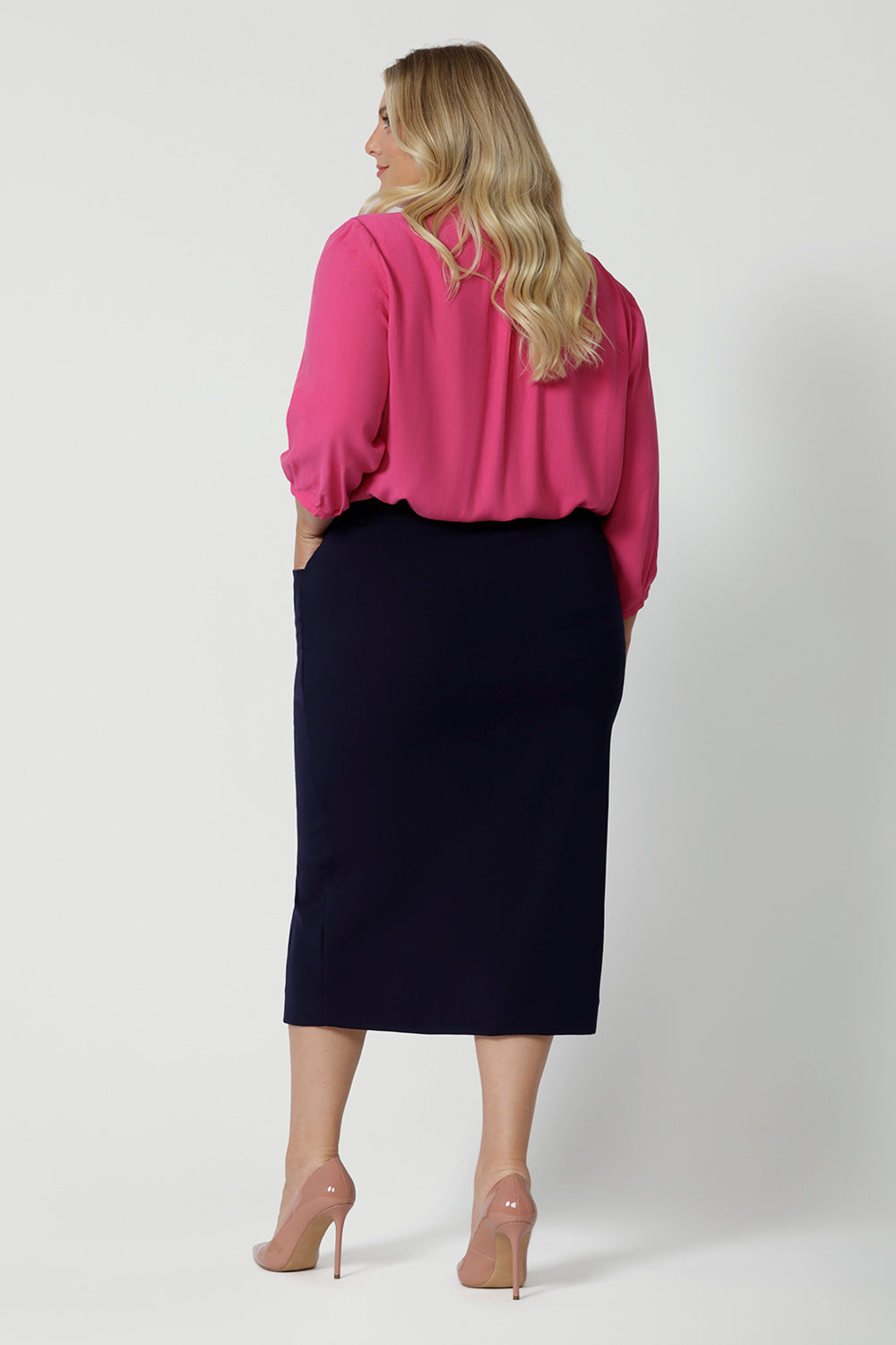 Full body back view of plus sized model wearing 3/4 length skirt. Work skirt is made of easy to care fabric in scube crepe fabric. Workwear skirt has a deep navy colour. This comfortable work wear skirt provides full body stretch great for any office or corporate setting. Australia made clothing. Made by Australian and New Zealand women. Leina and Fleur is a woman’s fashion label that stocks sizes from 8 to 24. Shop this appropriate work skirt in plus size and petite sizes. 