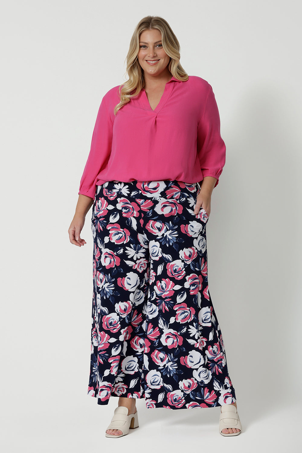 Full length body shot of plus sized woman wearing floral wide legged pants. The pant is designed with floral fabric combining white and pink on navy base. Easy care dry touch jersey fabric, pants designed in Australia by Australian and New Zealand women. Leina & Fleur is a fashion retailer that stock sizes from 8 to 24 with plus and petite sizes available. 