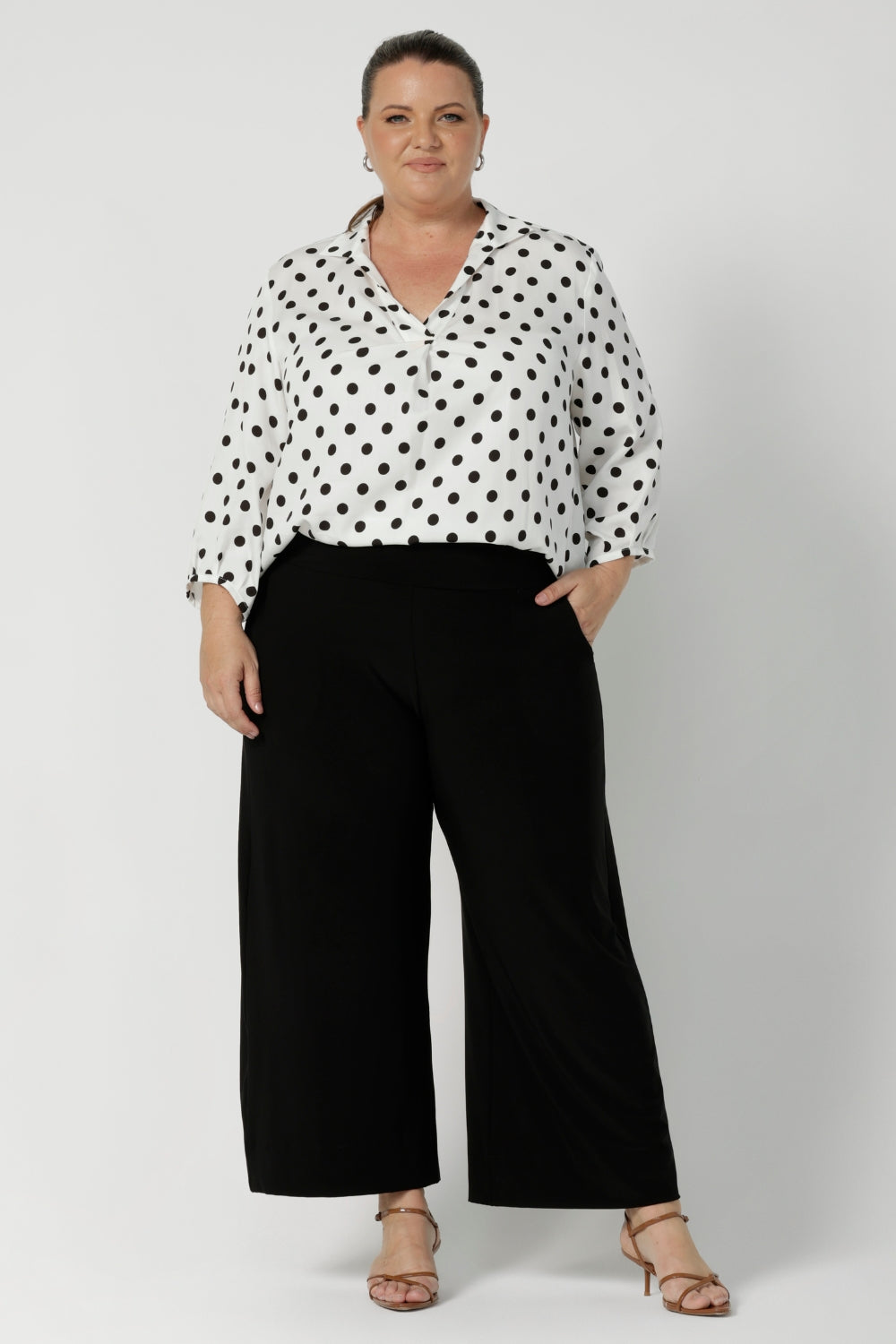 Plus sized woman wears white polka dotted blouse with 3/4-length sleeves, fixed collar and v neckline. Available in sizes 8-24, this versatile piece is proudly made in Australia, blending style with conscious design by Australian clothing brand Leina & Fleur.