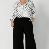 Plus sized woman wears white polka dotted blouse with 3/4-length sleeves, fixed collar and v neckline. Available in sizes 8-24, this versatile piece is proudly made in Australia, blending style with conscious design by Australian clothing brand Leina & Fleur.