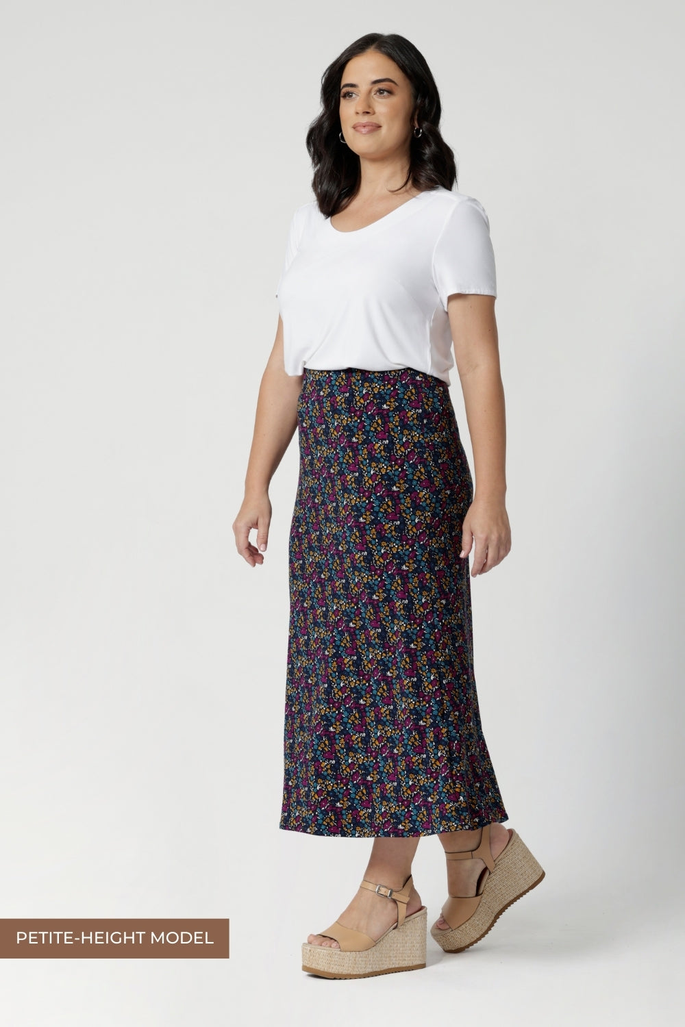 Side view image of petite height woman wearing floral midid skirt paired with white top and platform heels creating an easy care, easy wear summer outfit. Woman's clothing brand Leina & Fleur stock these midi skirts in sizes 8-24 with the options of 4 different colours & shades.