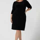 Size 18 woman wears the Audrey Dress, a black jersey round neckline dress with pockets and a boat neckline. Knee length style and easy care workwear for women. Made in Australia for women size 8 - 24. Styled back with a beige suede pump heel shoe. 