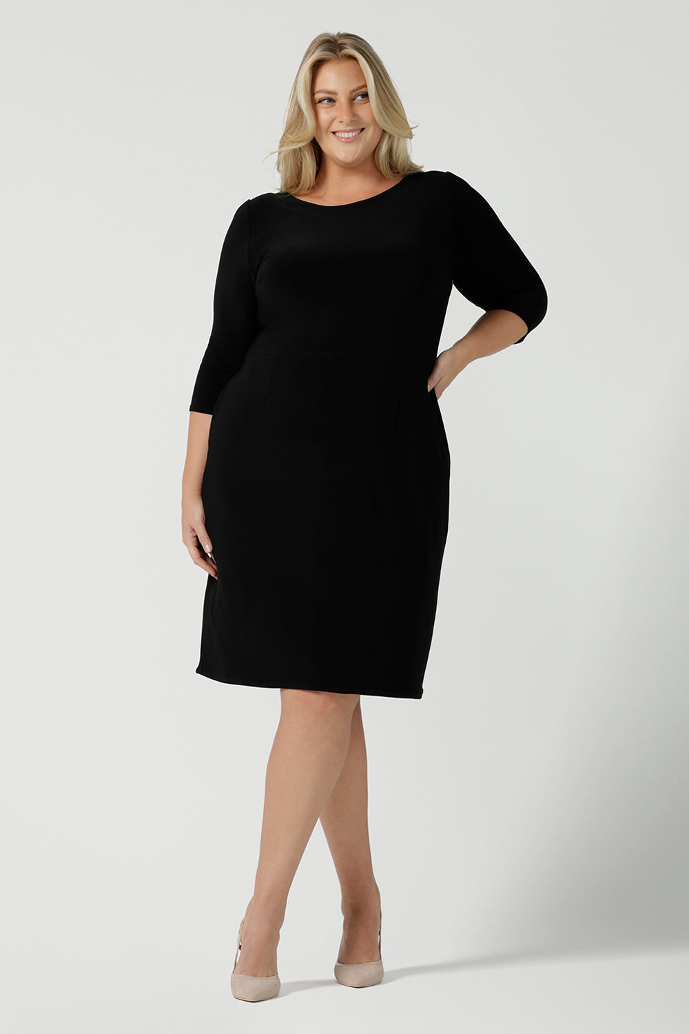 Size 18 woman wears the Audrey Dress, a black jersey round neckline dress with pockets and a boat neckline. Knee length style and easy care workwear for women. Made in Australia for women size 8 - 24. Styled back with a beige suede pump heel shoe. 