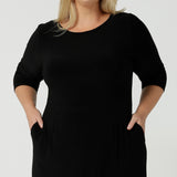 Close up of a size 18 woman wears the Audrey Dress, a black jersey round neckline dress with pockets and a boat neckline. Knee length style and easy care workwear for women. Made in Australia for women size 8 - 24. Styled back with a beige suede pump heel shoe.