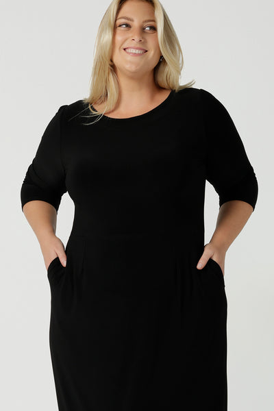 Close up of a size 18 woman wears the Audrey Dress, a black jersey round neckline dress with pockets and a boat neckline. Knee length style and easy care workwear for women. Made in Australia for women size 8 - 24. Styled back with a beige suede pump heel shoe.