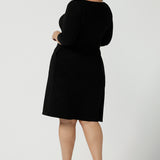 Back view of a size 18 woman wears the Audrey Dress, a black jersey round neckline dress with pockets and a boat neckline. Knee length style and easy care workwear for women. Made in Australia for women size 8 - 24. Styled back with a beige suede pump heel shoe.