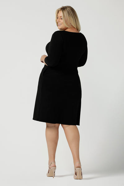 Back view of a size 18 woman wears the Audrey Dress, a black jersey round neckline dress with pockets and a boat neckline. Knee length style and easy care workwear for women. Made in Australia for women size 8 - 24. Styled back with a beige suede pump heel shoe.