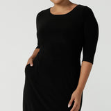 Close up of a size 10 woman wears the Audrey Dress, a black jersey round neckline dress with pockets and a boat neckline. Knee length style and easy care workwear for women. Made in Australia for women size 8 - 24.