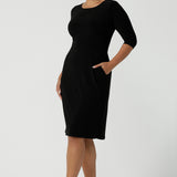 Size 10 woman wears the Audrey Dress, a black jersey round neckline dress with pockets and a boat neckline. Knee length style and easy care workwear for women. Made in Australia for women size 8 - 24. 