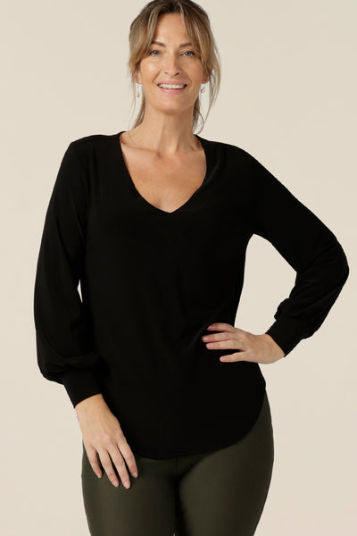 A size 10 woman wears a V-neck black top with long sleeves. This long sleeved top features cuffs at the wrists and a shirttail hemline. Made in Australia by Australian and New Zealand women's clothing label, L&F.