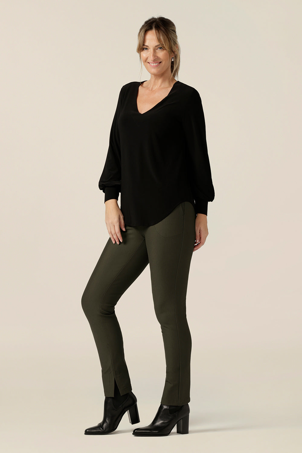 A size 10 woman wears a V-neck black top with long sleeves together with slim leg, olive green pants in stretch jersey. This long sleeved top features cuffs at the wrists and a shirttail hemline. Made in Australia by Australian and New Zealand women's clothing label, L&F.