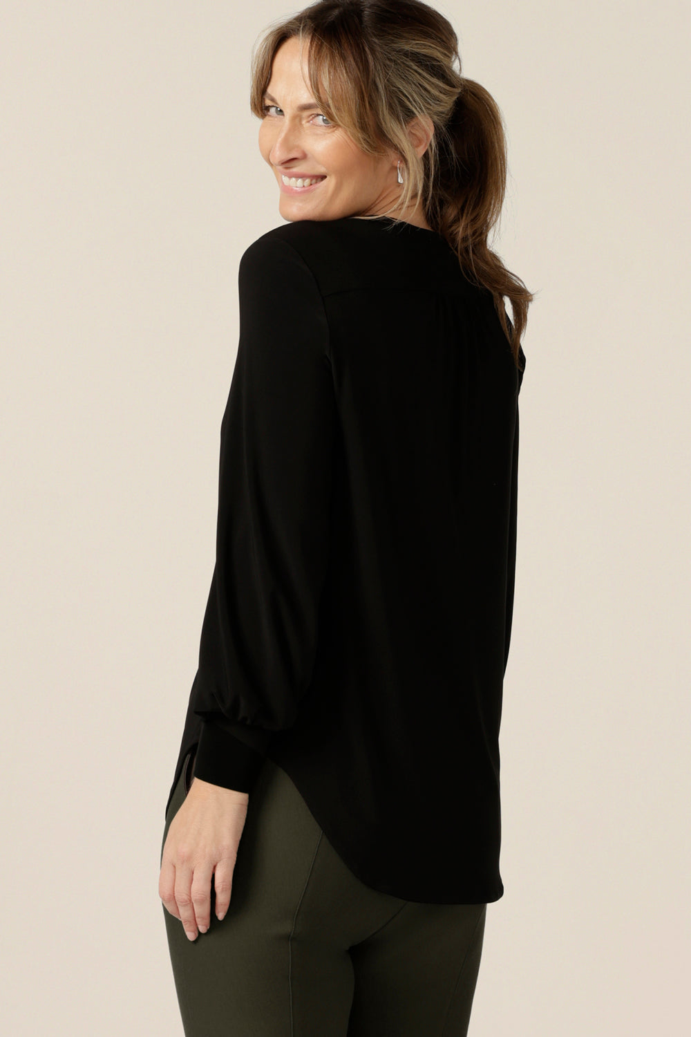 Back view of a size 10 woman wearing a V-neck black top with long sleeves. This long sleeved top features cuffs at the wrists and a shirttail hemline. Made in Australia by Australian and New Zealand women's clothing label, L&F.