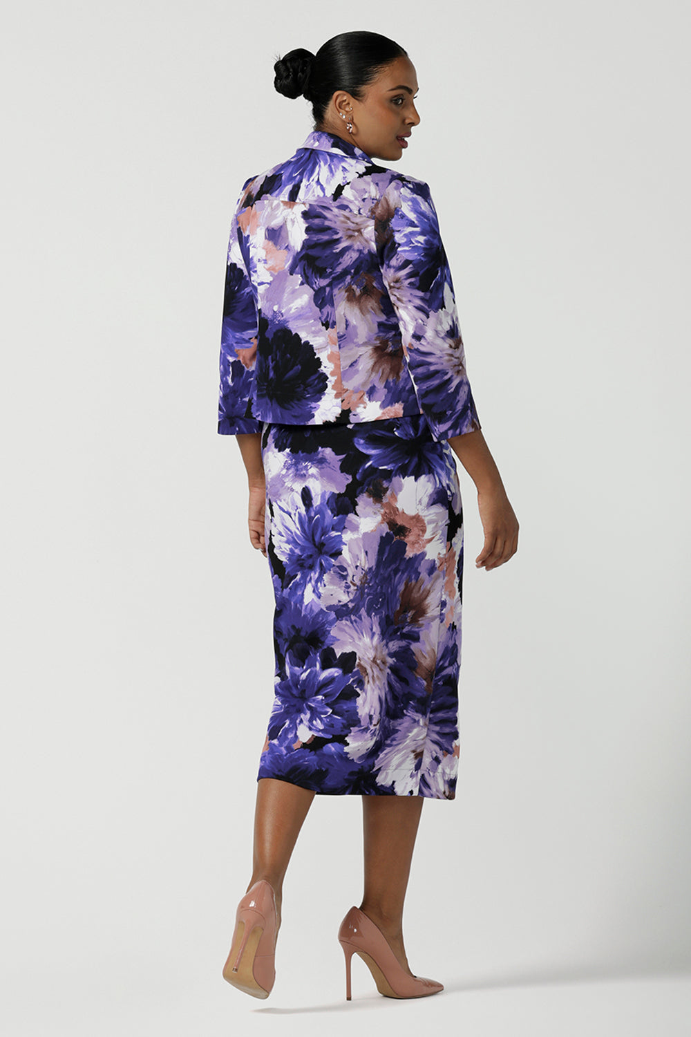 A size 10 woman wearing the Aaron Midi Skirt in Jacaranda. A printed Midi length skirt in purple florals. Made in Australia for women size 8-24. Styled back with pink pumps and a purple Jacaranda jacket.