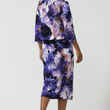 A size 10 woman wearing the Aaron Midi Skirt in Jacaranda. A printed Midi length skirt in purple florals. Made in Australia for women size 8-24. Styled back with pink pumps and a purple Jacaranda jacket.