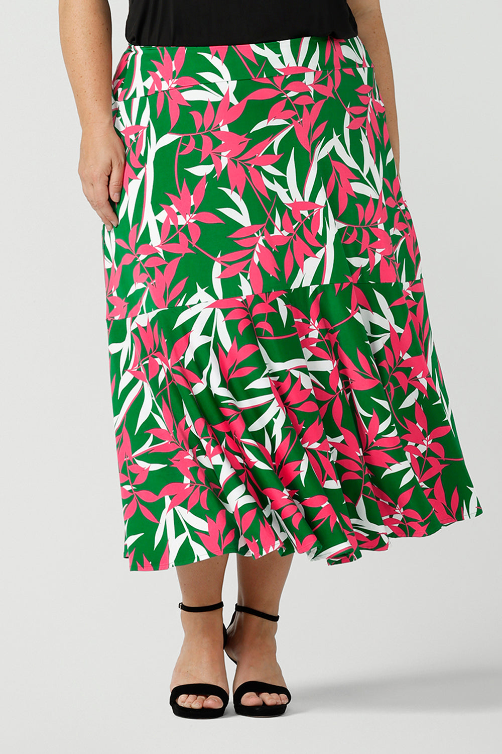 A size 12 woman wears a green jersey skirt with a leaf print. On a verdant green base in a soft jersey fabric. Styled back with black heels. Made in Australia for women size 8 - 24.