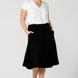 A good workwear skirt for women, this knee-length, pull-on black skirt has pockets, a ruffle hemline and comfort waistband. Worn with a V-neck, short sleeve top in white bamboo jersey, it's shown on a size 12 woman as a skirt for curvy office wear. A great addition to your capsule wardrobe, this black skirt is made by Australian made women's clothing brand, Leina & Fleur. Shop petite to plus size skirts in their online boutique in Australia. 