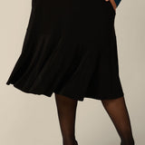 A fuller figure woman wears a pull on, black skirt with knee-length ruffle hemline and side pockets. This black skirt is a comfortable skirt for workwear or smart-casual wear.