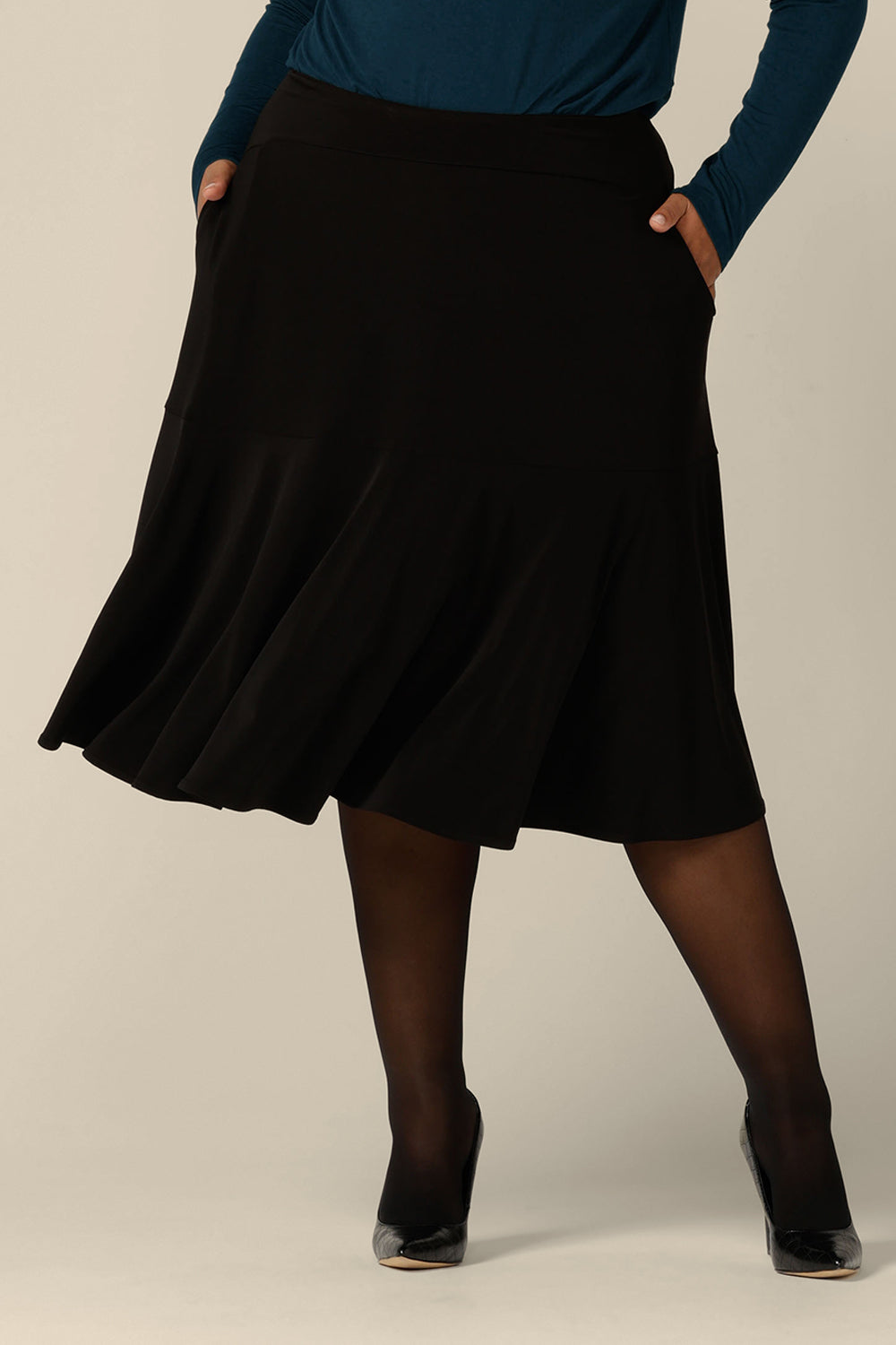 A fuller figure woman wears a pull on, black skirt with knee-length ruffle hemline and side pockets. This black skirt is a comfortable skirt for workwear or smart-casual wear.