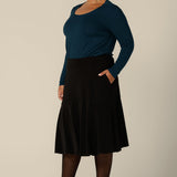 A plus size woman wears a pull on, black skirt with knee-length ruffle hemline and side pockets. This black skirt is a comfortable skirt for workwear or smart-casual wear.