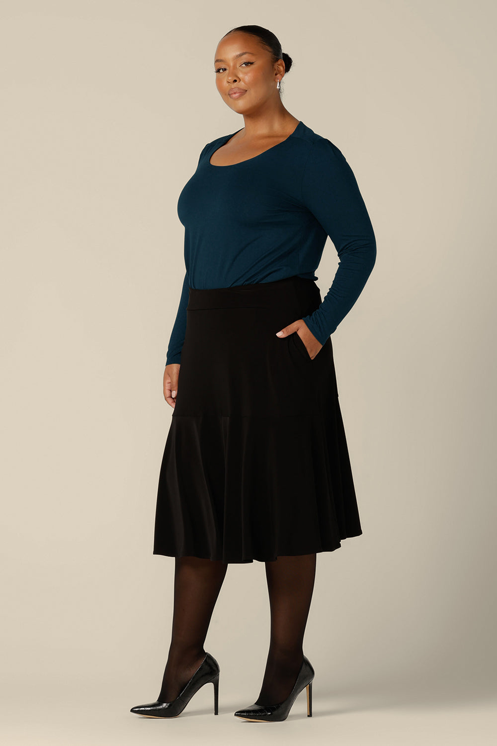 A plus size woman wears a pull on, black skirt with knee-length ruffle hemline and side pockets. This black skirt is a comfortable skirt for workwear or smart-casual wear.
