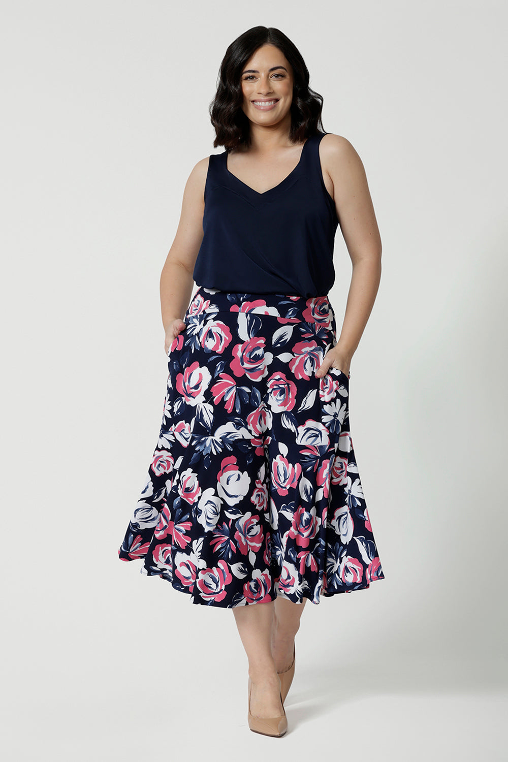 Full body close up shot of petite woman wearing three quarter skirt that falls to her mid calf. Printed floral fabric showcases white and pink roses on a navy base. This easy to care fabric is used to craft garment in Australia designed by Australian and New Zealand women. Leina & Fleur proudly stocks petite and plus sizes ranging from 8-24.