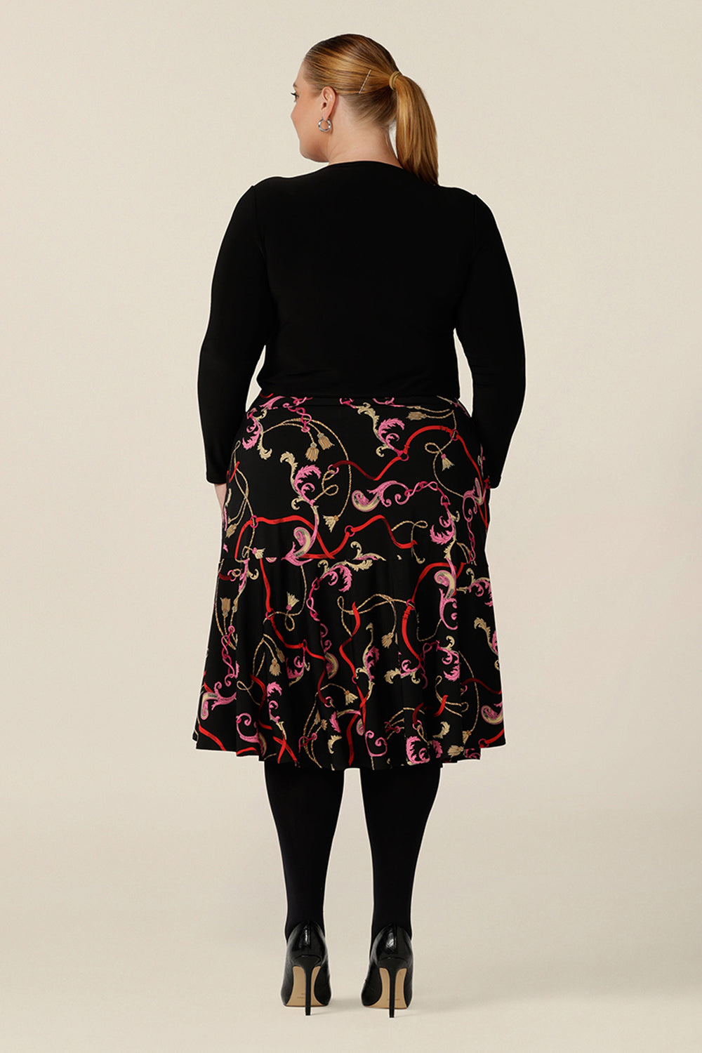 Back view of a curvy, size 18 woman wearing a good skirt for plus size workwear. The Berit Skirt in Tassel is a pull-on knee length skirt with pockets and ruffle hem, and is worn with a square neck, long sleeve black top. Both women's plus size skirt and top are made in Australia by Australian fashion brand, L&F.