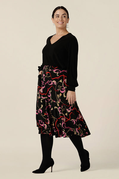 A woman wears a printed skirt for work. The Berit Skirt in Tassel is a pull-on knee length skirt with pockets and ruffle hem, and is worn with a V neck, long bishop sleeve black top. Both women's plus size skirt and top are made in Australia by Australian fashion brand, L&F.