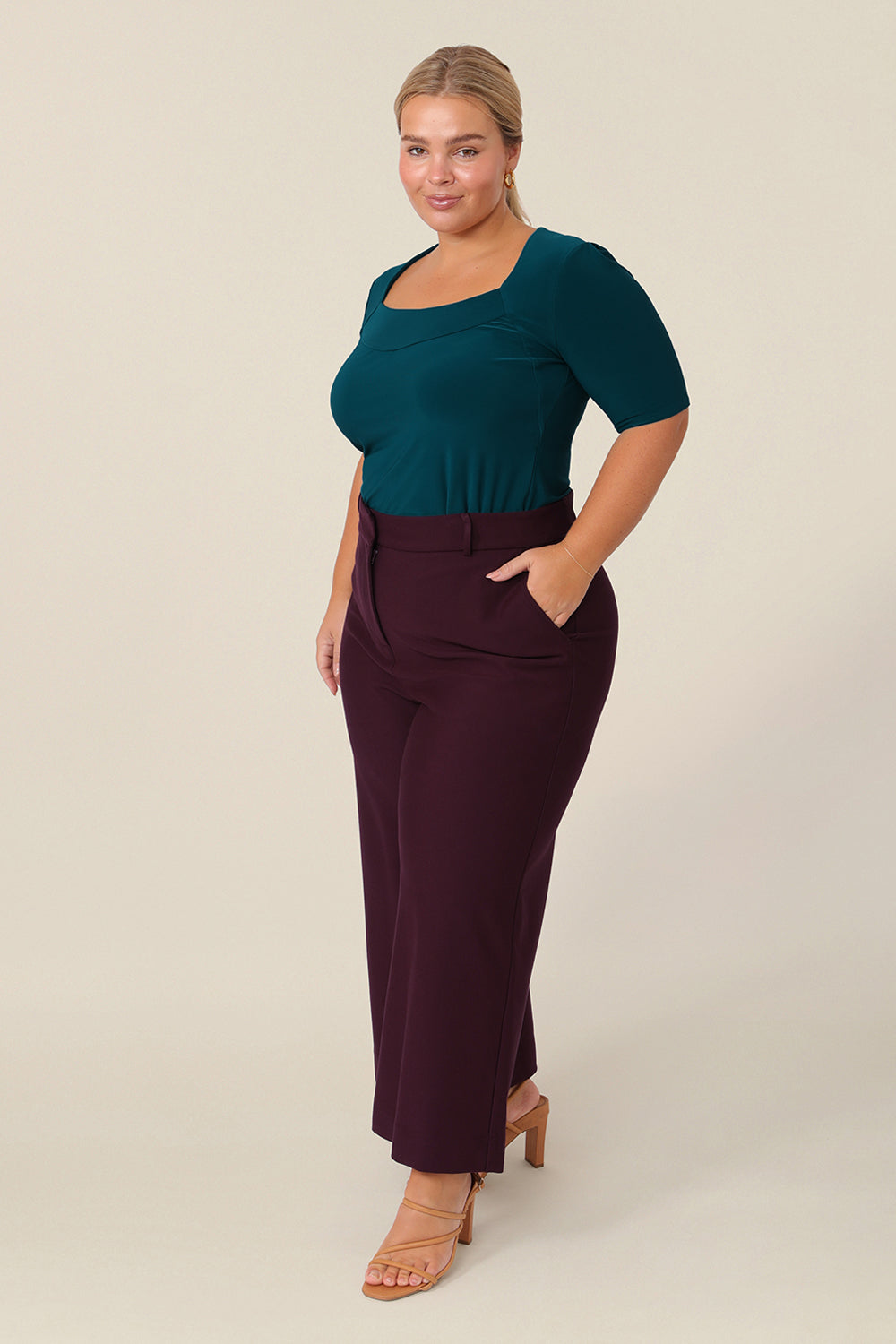 A size 14 curvy woman wears the short sleeve Berni Top in dark teal stretch jersey. The top has a squared scoop neckline, a structured bust and short sleeves. The top is paired with tailored wide leg pants in Mulberry for a work wear look. Made in Australia, buy women's jersey tops in sizes 8 to size 24 online at Leina and Fleur.