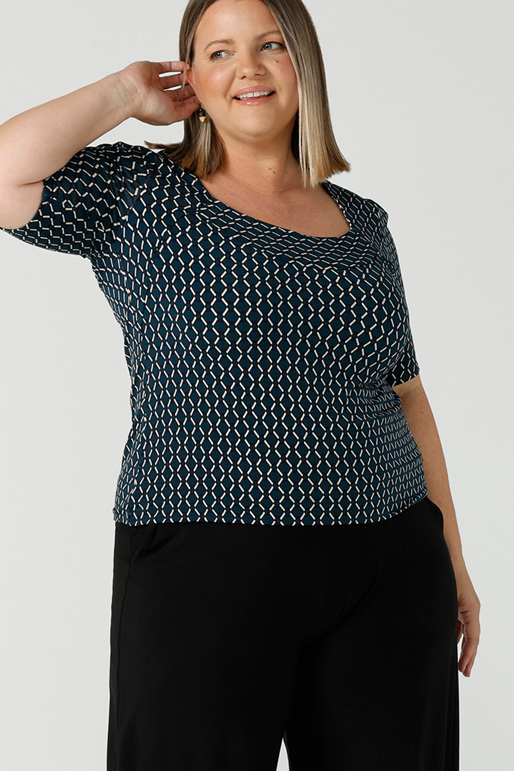 A plus size 16 woman wears the Berni Top in Infinity with a jade base colour and geometric pattern. A comfortable jersey work top style back with the Lulu pants in navy. Made in Australia for women size 8 - 24.