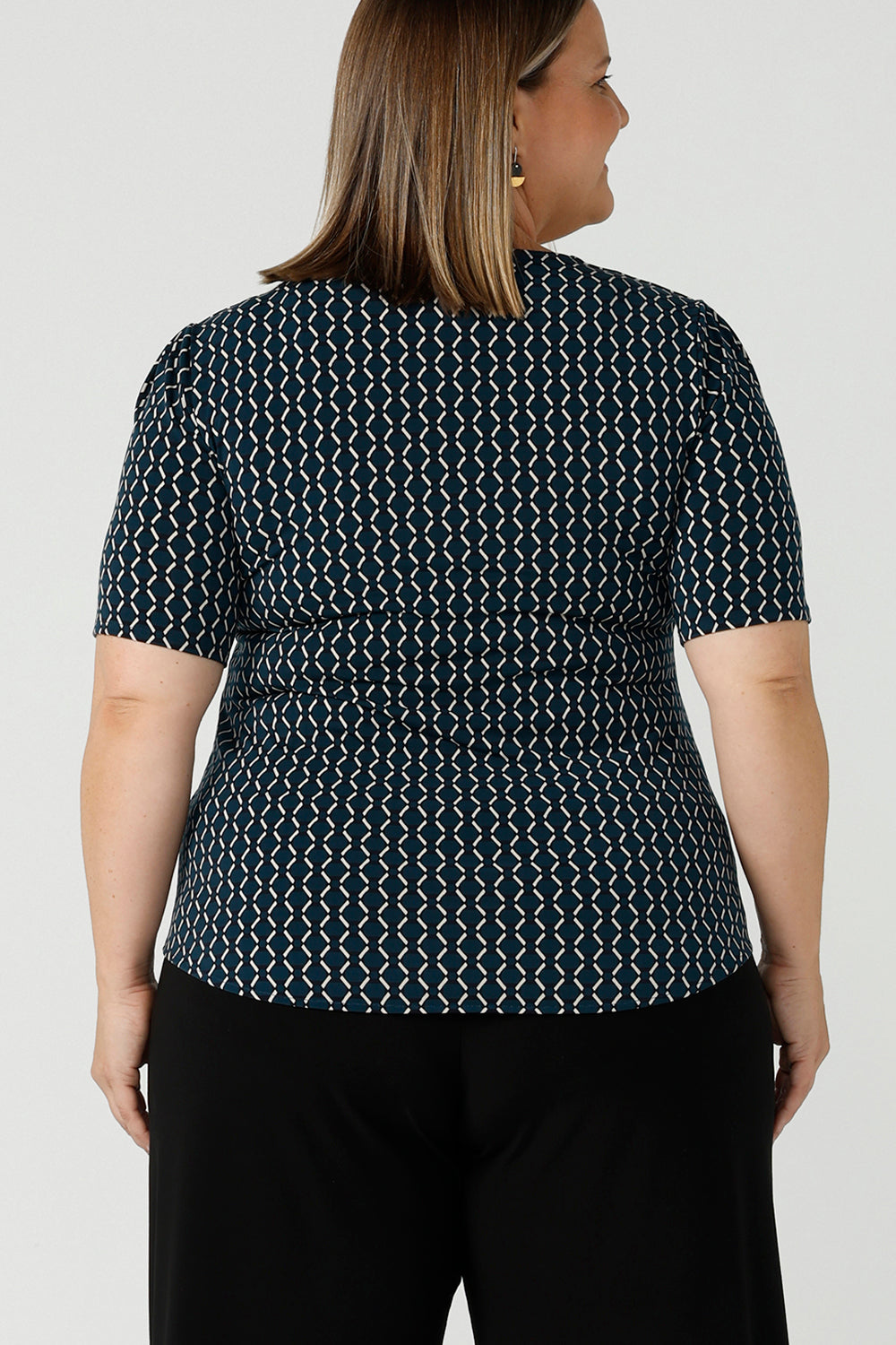Back view of a size 16 woman wears the Berni Top in Infinity with a jade base colour and geometric pattern. A comfortable jersey work top style back with a wide leg Bradley pant in black. Made in Australia for women size 8 - 24.