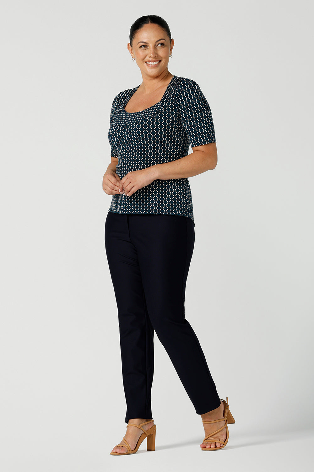 A size 12 woman wears the Berni Top in Infinity with a jade base colour and geometric pattern. A comfortable jersey work top style back with the Lulu pants in navy. Made in Australia for women size 8 - 24.