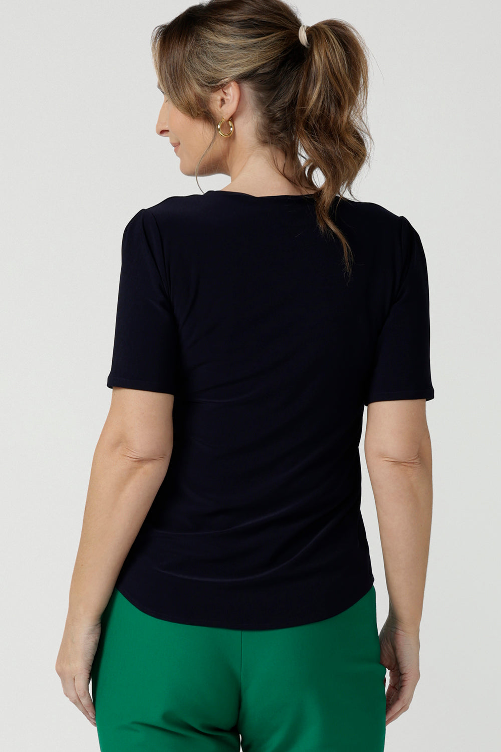 Back view of A size 10 over 40 year old woman wears a casual jersey top in navy blue, styled with emerald green pants. This Australian-made women's top has short fitted sleeves, a square neckline and and high-low - perfect for weekend casual and travel capsule wardrobes.