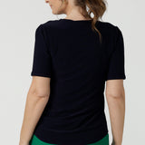 Back view of A size 10 over 40 year old woman wears a casual jersey top in navy blue, styled with emerald green pants. This Australian-made women's top has short fitted sleeves, a square neckline and and high-low - perfect for weekend casual and travel capsule wardrobes.