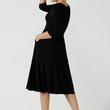 Back view of a size 10 woman wears the Bettina dress in black soft jersey. Wrap front reversible design with pockets and a tier. Styled back with black sling back shoes. Made in Australia for women size 8 - 24.