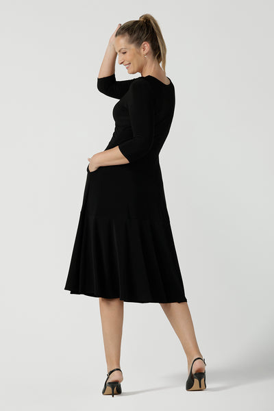 Back view of a size 10 woman wears the Bettina dress in black soft jersey. Wrap front reversible design with pockets and a tier. Styled back with black sling back shoes. Made in Australia for women size 8 - 24.
