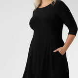 Size 18 woman wears a black reversible Bettina dress in black Jersey dress made in Australia. V-neck style with pockets and midi length. Made in Australia for women size 8 - 24. Reversible dress can be worn featuring boat neck or neck.