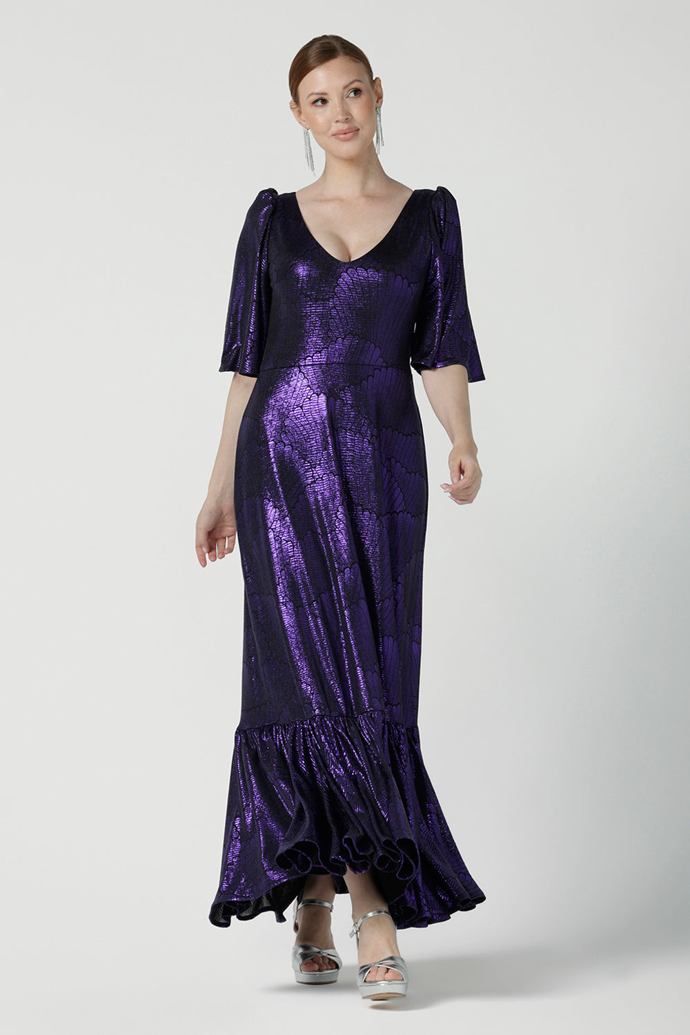 A size 10 woman wears a glamorous maxi dress with a ruffle hem and low v-neckline. Featuring a flutter sleeve and made in shiny purple jersey with a foil print. Made in Australia for women for evening gown cocktail wear. Size 8 - 24.