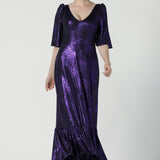 A size 10 woman wears a glamorous maxi dress with a ruffle hem and low v-neckline. Featuring a flutter sleeve and made in shiny purple jersey with a foil print. Made in Australia for women for evening gown cocktail wear. Size 8 - 24.