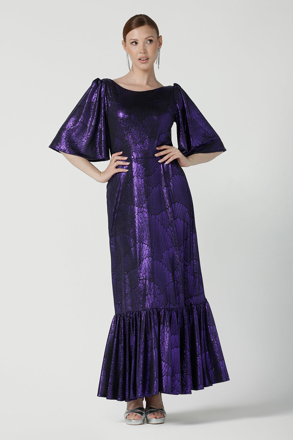 A size 10 woman wears a glamorous maxi dress with a ruffle hem and low v-neckline. Featuring a flutter sleeve and made in shiny purple jersey with a foil print. Made in Australia for women for evening gown cocktail wear. Size 8 - 24.