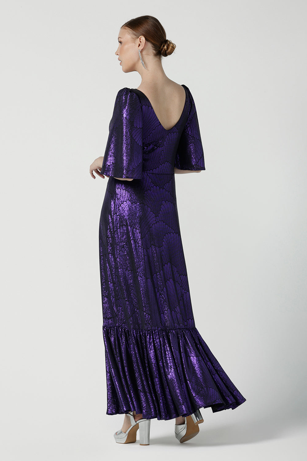 A size 10 woman wears a glamorous maxi dress with a ruffle hem and low v-neckline. Featuring a flutter sleeve and made in shiny purple jersey with a foil print. Made in Australia for women for evening gown cocktail wear. Size 8 - 24.