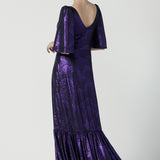 A size 10 woman wears a glamorous maxi dress with a ruffle hem and low v-neckline. Featuring a flutter sleeve and made in shiny purple jersey with a foil print. Made in Australia for women for evening gown cocktail wear. Size 8 - 24.