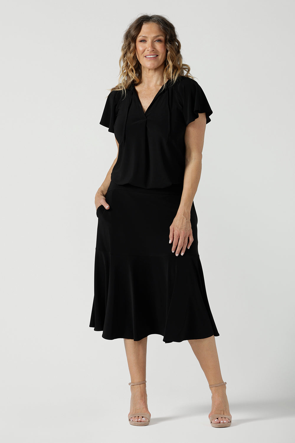 A size 10 woman wears the Bowie Top in Black. A jersey flutter sleeve style with a v-neckline and tie neck. Made in Australia for women 8-24.